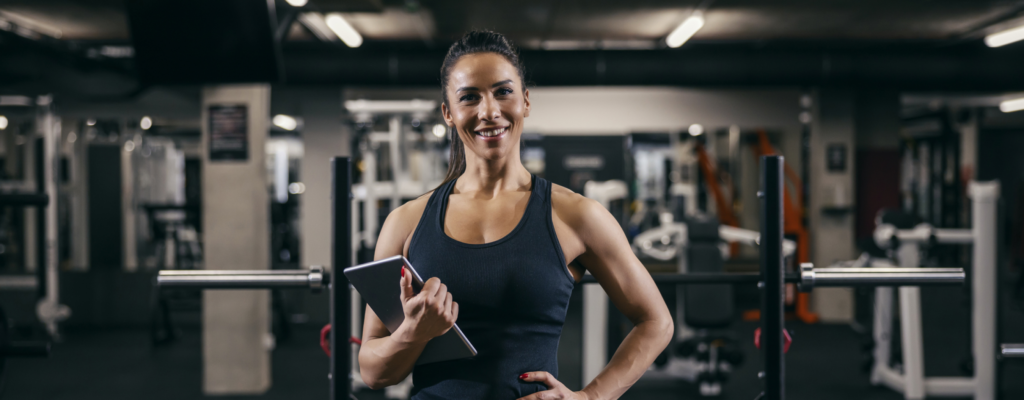 Female coach in gym holding a tablet. She is confident and smiling.